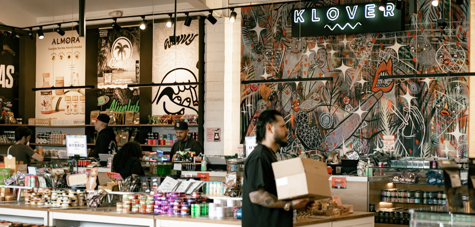 Inside Klover dispensary — counter scene under the KLOVER neon sign with the WAVVY × KLOVER mural wall
