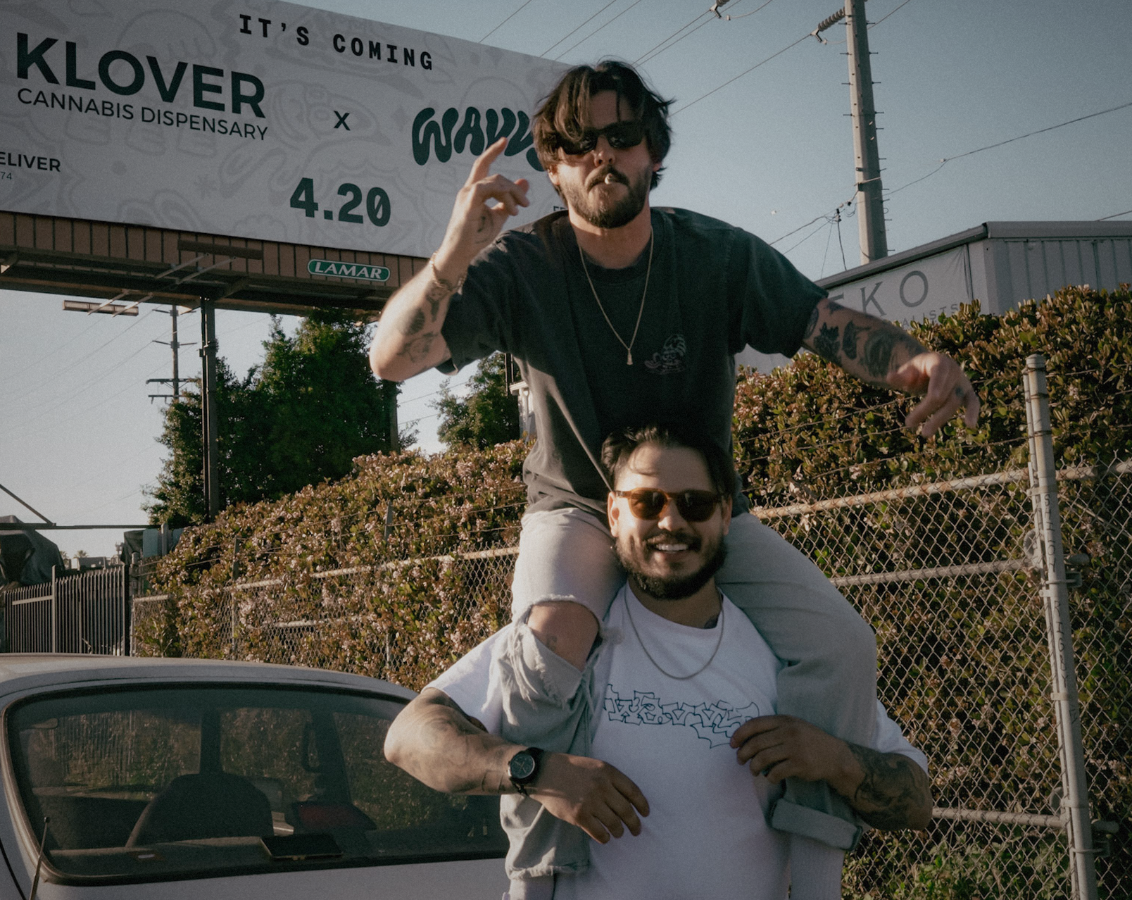 Two Wavvy crew under the WAVVY × KLOVER billboard at dusk